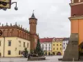 Rynek w Tarnowie zimą, z choinką i ludźmi przy fontannie. W tle zabytkowe kamienice i wieża ratuszowa.