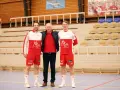 Damian Kędziora z dwoma zawodnikami w strojach sportowych, na tle trybun hali sportowej.