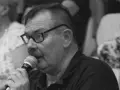 Krzysztof Janicki w okularach i z wąsami mówi do mikrofonu, w tle widać ludzi.