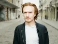 Bartosz Bednarski na tle miejskiej ulicy, z rudymi włosami i zielonymi oczami, w białej koszuli i ciemnej kurtce.