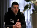 Maciej Pilitowski w czarnej bluzie z orłem i logiem Suzuki, z dłońmi złożonymi na stole, w tle zasłony i rośliny.