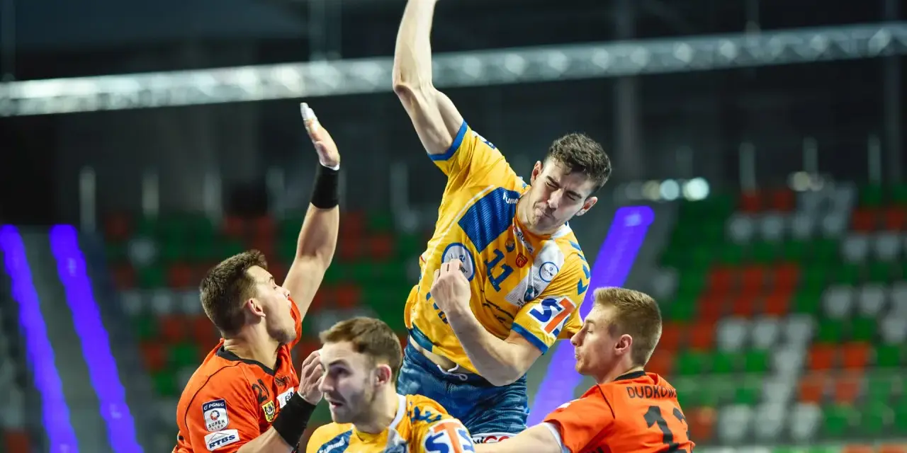Gracz w żółtej koszulce rzuca piłkę do bramki, broniony przez zawodników w pomarańczowych strojach. Emocjonujący moment z meczu final four handball.