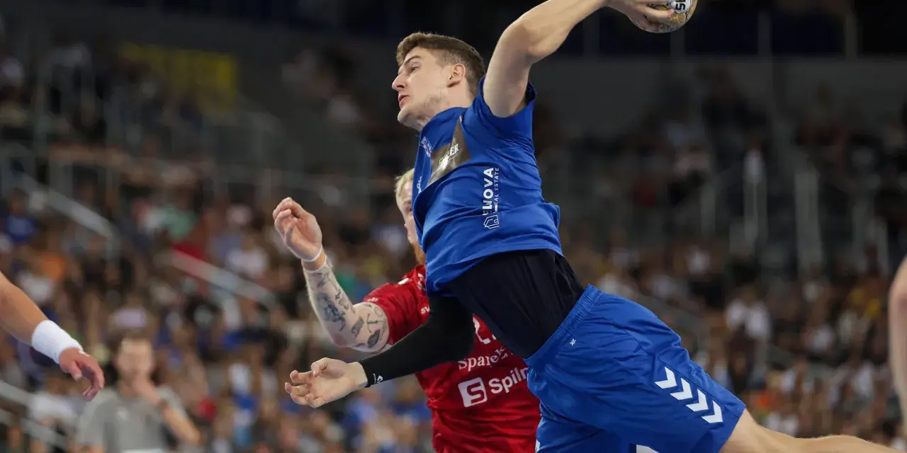 Magnus Landin Jacobsen w skoku do rzutu piłką ręczną, w niebieskim stroju, z widownią w tle.