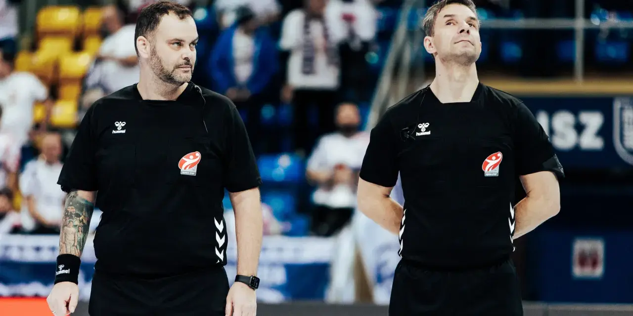 Dwóch sędziów w czarnych koszulkach z logo Hummel i emblematem związku sportowego, gotowych do rozpoczęcia meczu ligi piłki ręcznej.