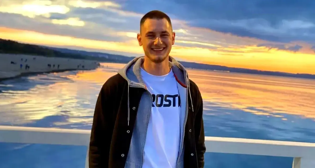 Uśmiechnięty Krzysztof Komarzewski na molo o zachodzie słońca. W tle plaża i morze.