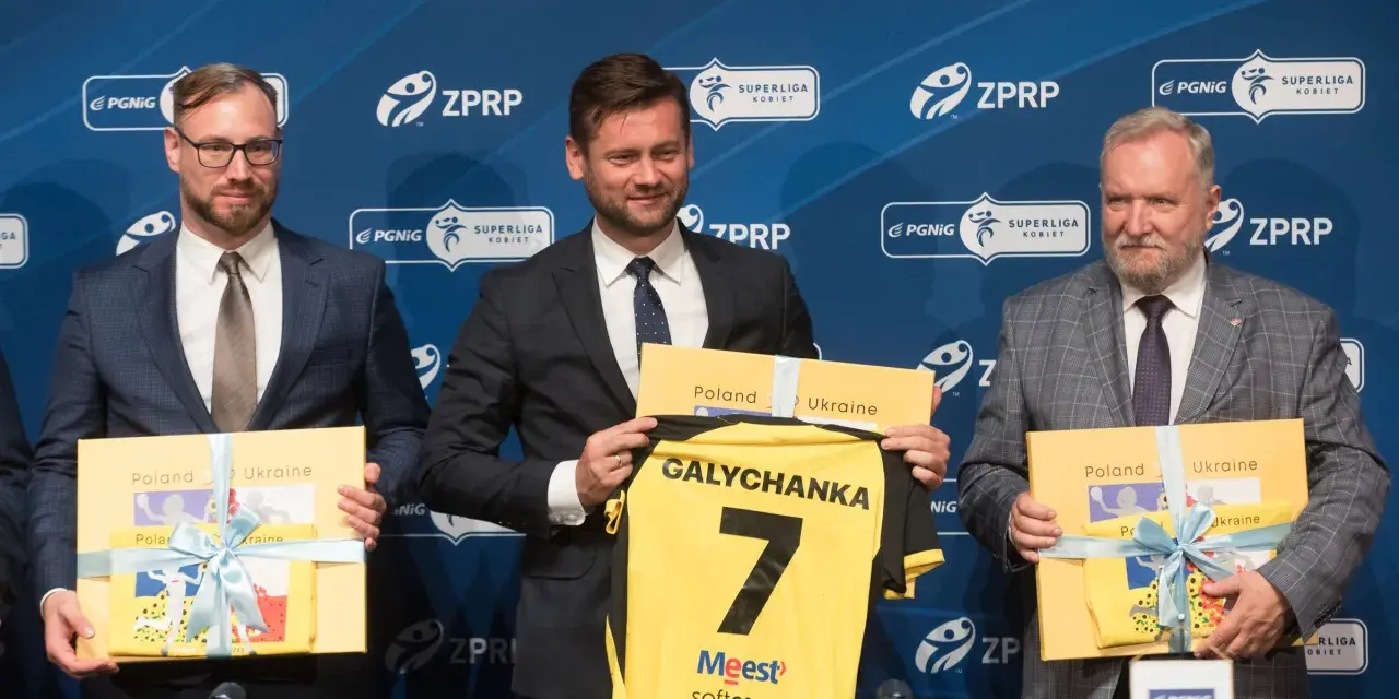 Trzech mężczyzn w garniturach prezentuje koszulkę z napisem "Galychanka" i numerem 7. Obok nich żółte pudełka z napisami "Poland Ukraine".