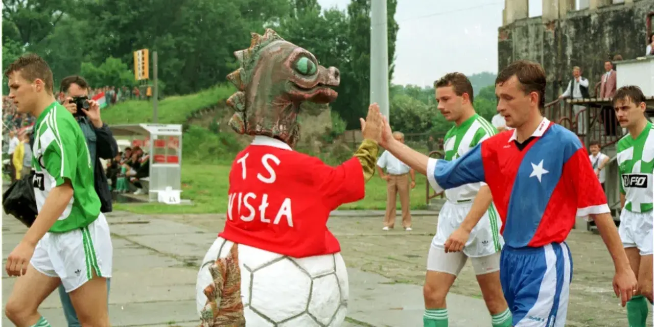 Artur Woźniak w barwach Wisły daje piątkę z maskotką. Piłkarze w zielonych strojach wychodzą na boisko.