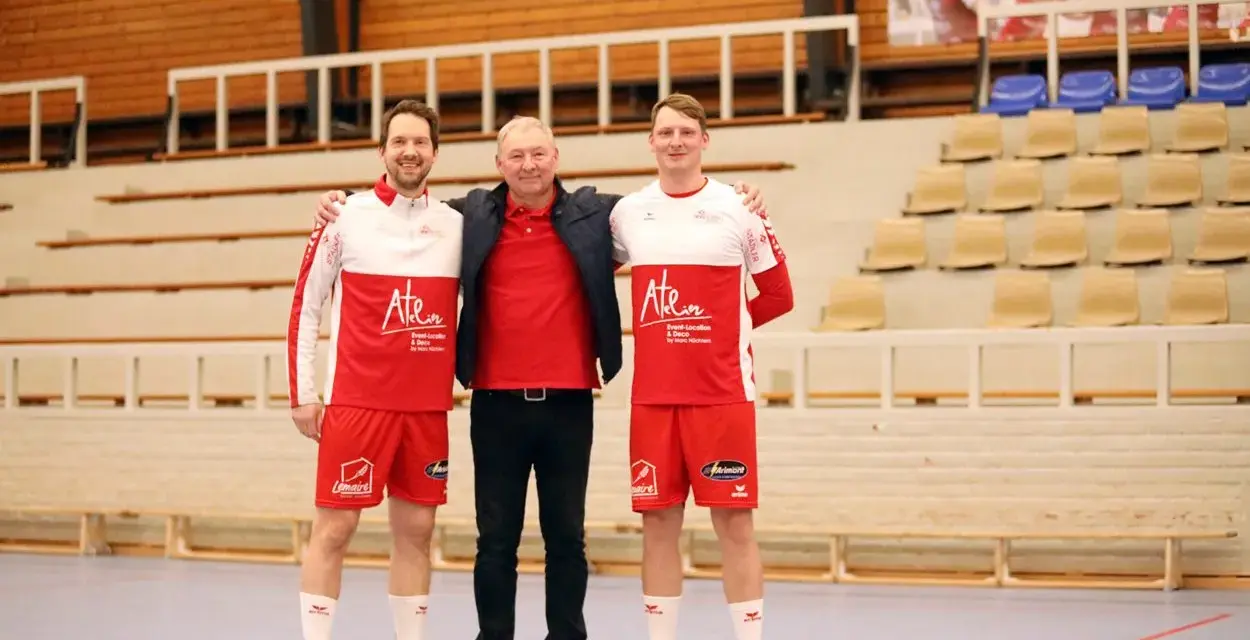 Damian Kędziora z dwoma zawodnikami w strojach sportowych, na tle trybun hali sportowej.