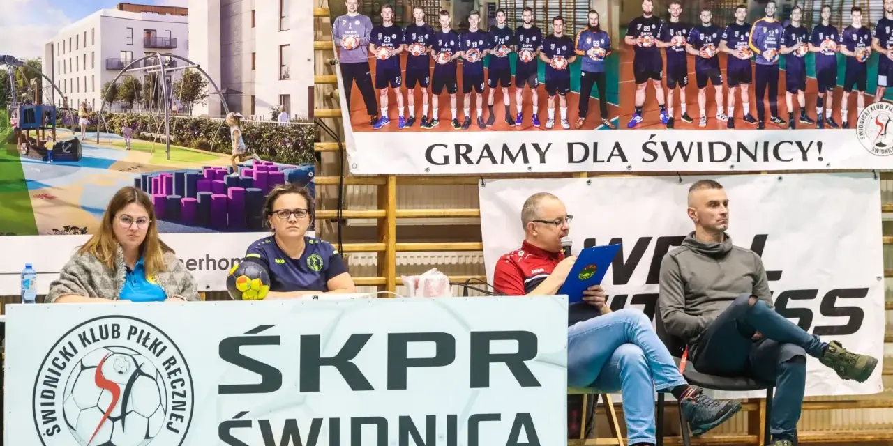 Na banerze widnieje drużyna SKPR Świdnica. Dwie kobiety siedzą za stołem z piłką ręczną.
