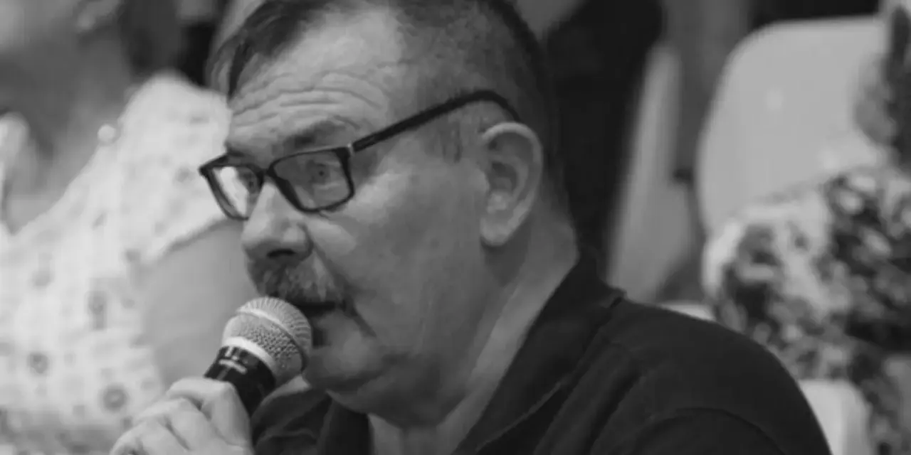 Krzysztof Janicki w okularach i z wąsami mówi do mikrofonu, w tle widać ludzi.