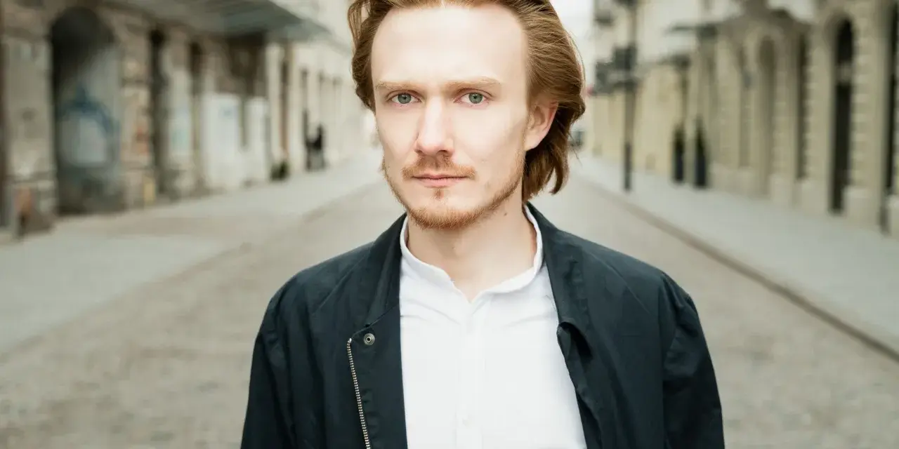 Bartosz Bednarski na tle miejskiej ulicy, z rudymi włosami i zielonymi oczami, w białej koszuli i ciemnej kurtce.