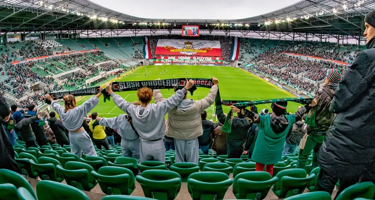 Gwardia Opole kibicuje na stadionie, trzymając szaliki. Na boisku trwa mecz, a trybuny wypełniają fani.