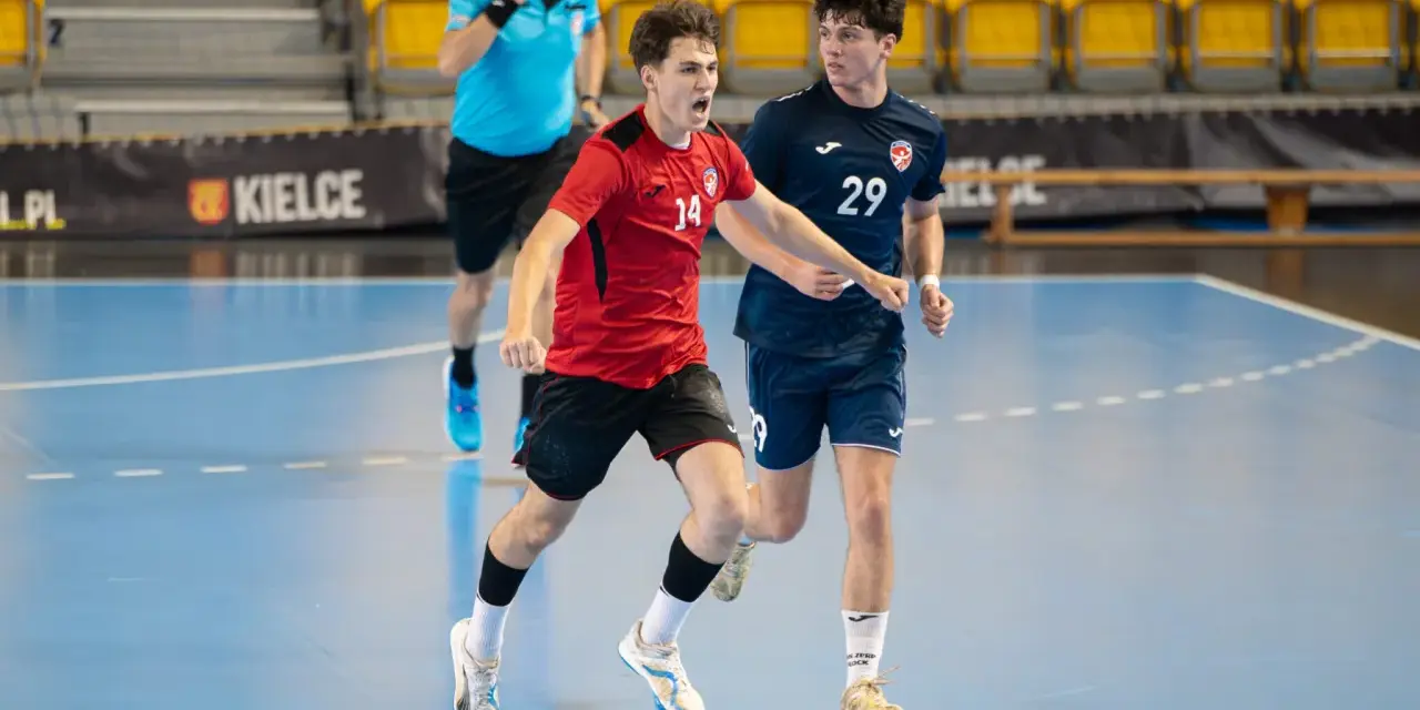 Jakub Smoliński w czerwonej koszulce z numerem 14 biegnie z piłką ręczną, obok zawodnik w granatowej koszulce z numerem 29.