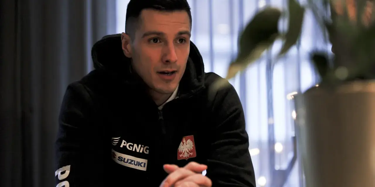 Maciej Pilitowski w czarnej bluzie z orłem i logiem Suzuki, z dłońmi złożonymi na stole, w tle zasłony i rośliny.