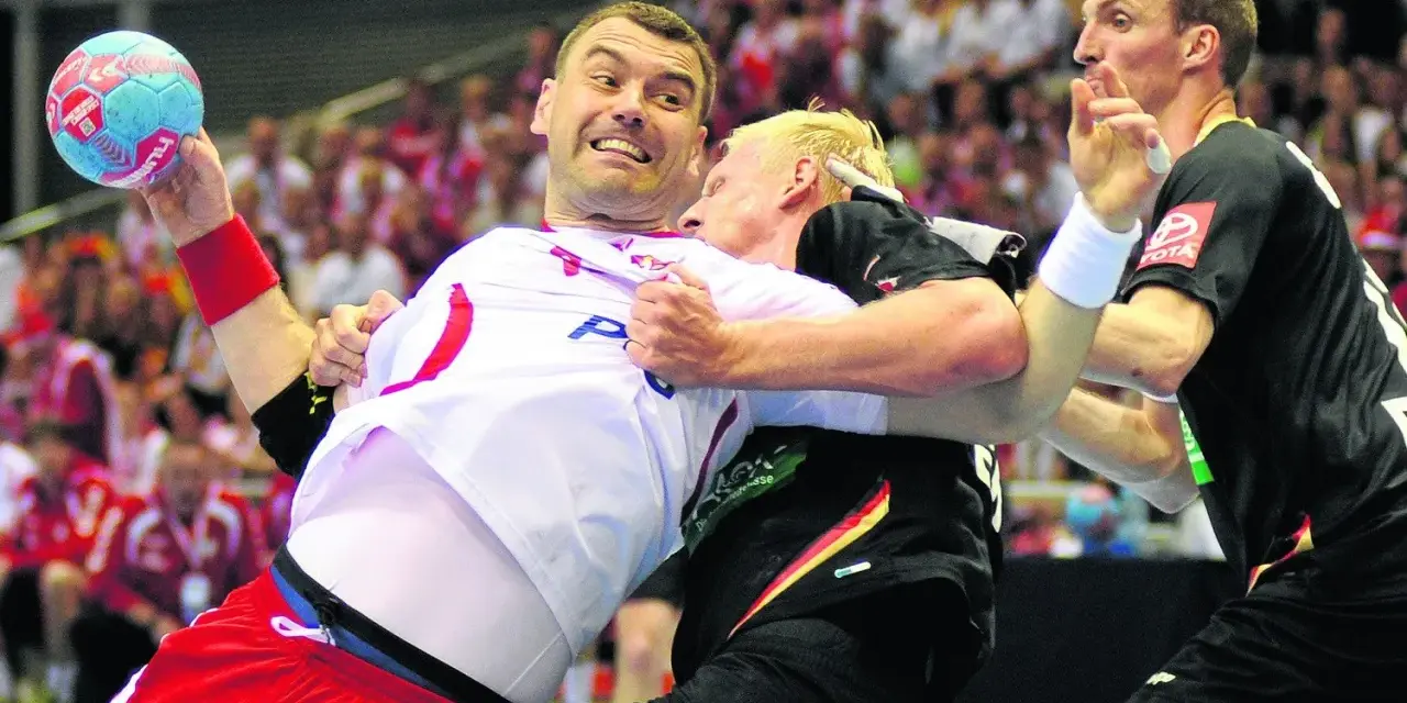 Gracz w białej koszulce z piłką ręczną próbuje wykonać rzut karny, podczas gdy przeciwnik próbuje go zatrzymać.
