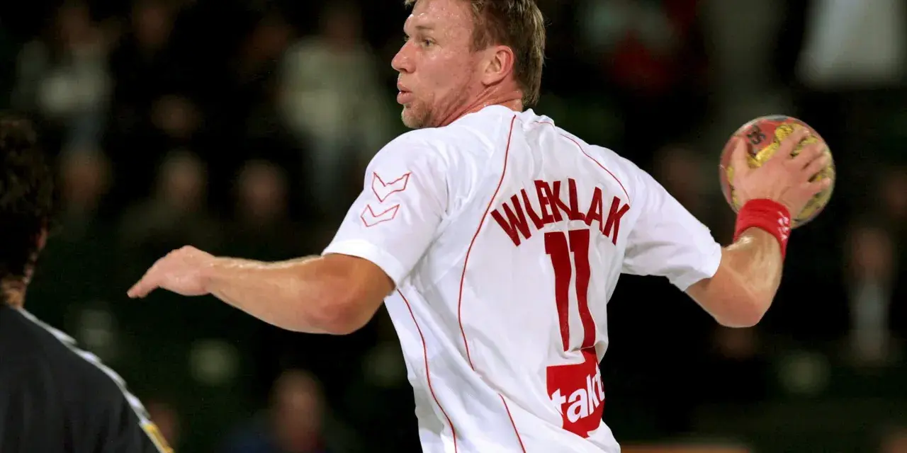 Damian Wleklak, numer 11, w białej koszulce z czerwonymi detalami, przygotowuje się do rzutu piłką ręczną.