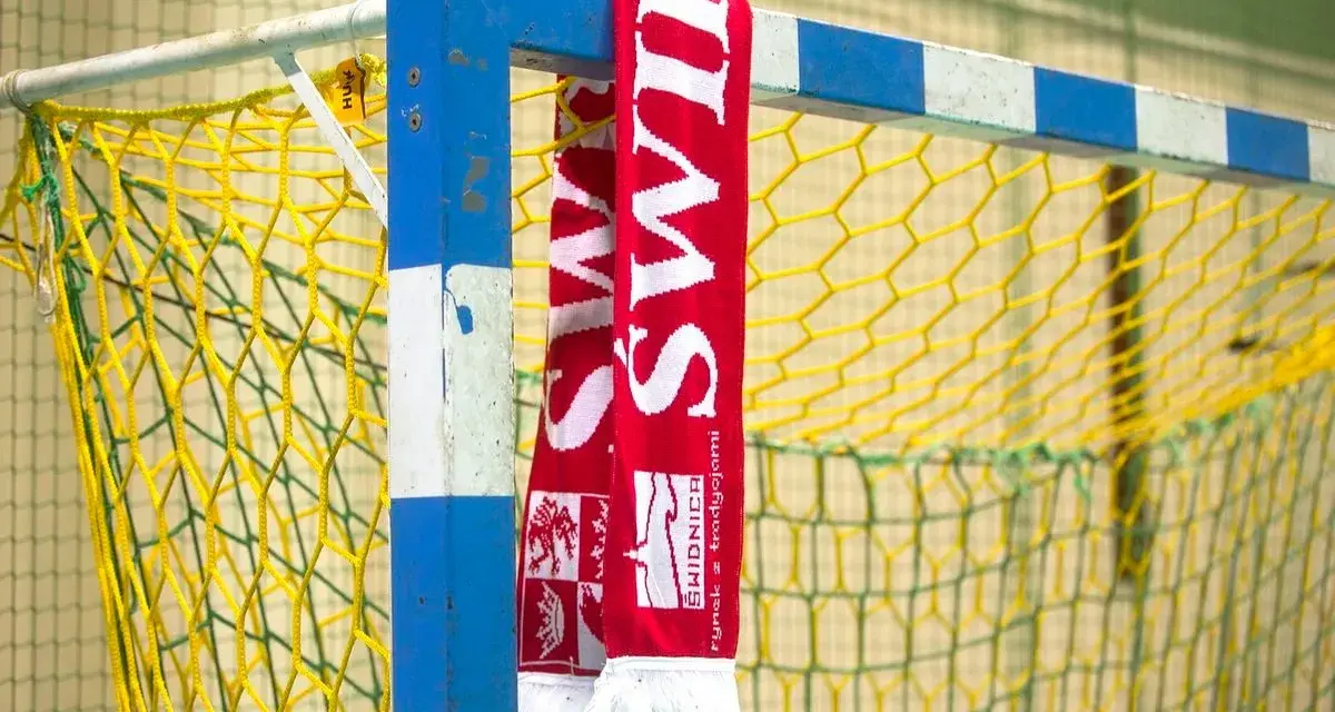Czerwono-biały szalik z napisem "ŚWIDNICA" wisi na niebiesko-białej bramce do piłki ręcznej z żółtą siatką.