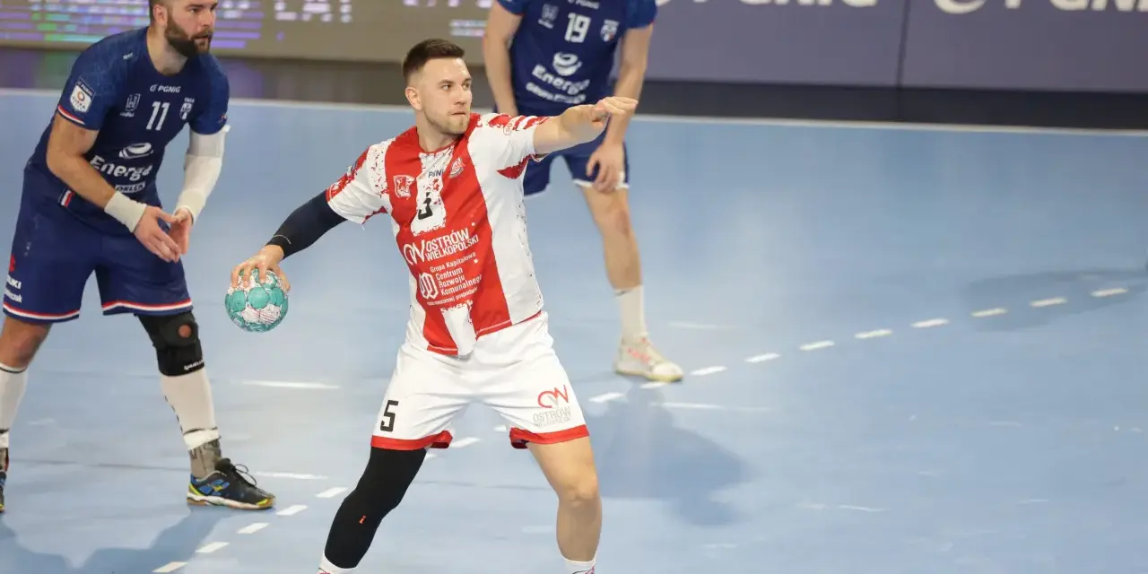 Kamil Adamski w akcji, rzuca piłkę do bramki. Zawodnik w białej koszulce z czerwonymi pasami i numerem 5.