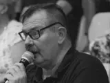 Krzysztof Janicki w okularach i z wąsami mówi do mikrofonu, w tle widać ludzi.