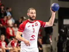 Gracz w białej koszulce z czerwonymi rękawami, numerem 3 i godłem Polski, trzyma niebieską piłkę ręczną, gotowy do rzutu.