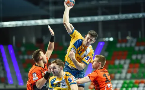 Gracz w żółtej koszulce rzuca piłkę do bramki, broniony przez zawodników w pomarańczowych strojach. Emocjonujący moment z meczu final four handball.