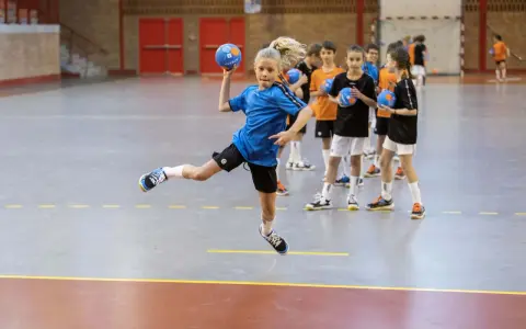 Dziewczynka w skoku wykonuje kozłowanie w piłce ręcznej, podczas gdy inni gracze czekają na swoją kolej.