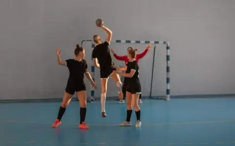 Piłkarki ręczne w akcji. Jedna z nich skacze do rzutu, podczas gdy koleżanki z drużyny i przeciwniczka obserwują. To dynamiczna gra pasywna w piłce ręcznej.