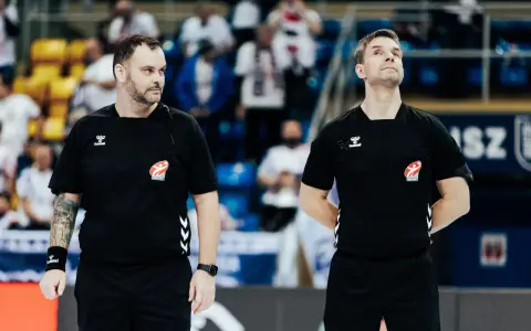 Dwóch sędziów w czarnych koszulkach z logo Hummel i emblematem związku sportowego, gotowych do rozpoczęcia meczu ligi piłki ręcznej.