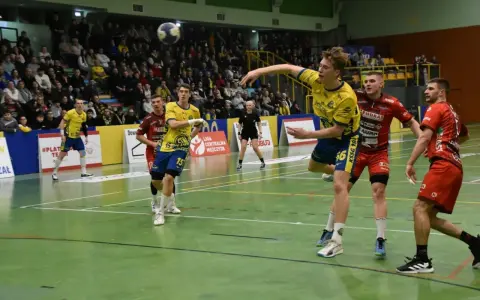 Gracz w żółtej koszulce rzuca piłkę ręczną podczas meczu. W tle kibice i zawodnicy w czerwonych strojach. Stal Gorzów walczy o zwycięstwo.