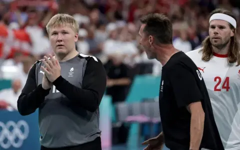 Trzech zawodników Danii w piłce ręcznej na boisku. Jeden z nich, w szarej koszulce, klaszcze.