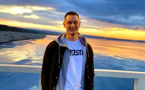 Uśmiechnięty Krzysztof Komarzewski na molo o zachodzie słońca. W tle plaża i morze.