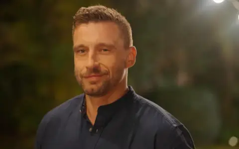 Uśmiechnięty Jakub Wojnowski w granatowej koszuli, w tle rozmyte światła i zieleń.