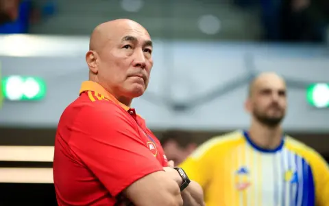Skupiony trener Vive Kielce w czerwonej koszulce z pomarańczowym kołnierzem, z rękami skrzyżowanymi na piersi.