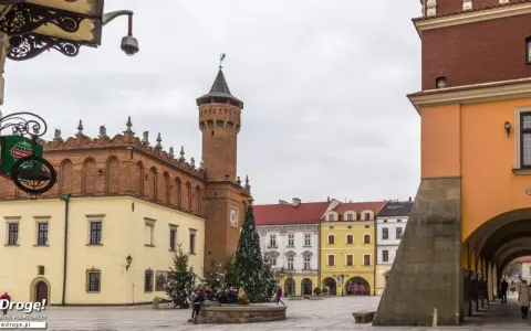 Rynek w Tarnowie zimą, z choinką i ludźmi przy fontannie. W tle zabytkowe kamienice i wieża ratuszowa.