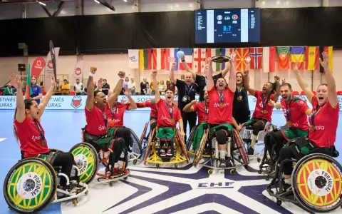 Drużyna Portugalii w piłce ręcznej na wózkach świętuje zwycięstwo. Na ekranie wynik meczu, a w tle flagi państwowe i logo EHF.