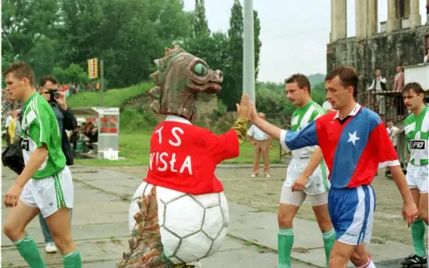 Artur Woźniak w barwach Wisły daje piątkę z maskotką. Piłkarze w zielonych strojach wychodzą na boisko.