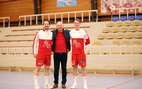 Damian Kędziora z dwoma zawodnikami w strojach sportowych, na tle trybun hali sportowej.