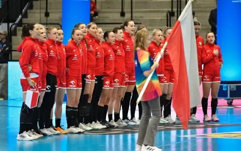 Drużyna piłki ręcznej kobiet Polski w czerwonych strojach stoi w rzędzie, trzymając polską flagę.