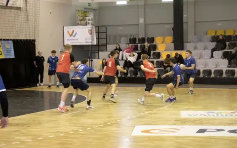 Kacper Wachowiak w akcji podczas meczu piłki ręcznej. Zawodnicy w czerwonych i niebieskich strojach walczą o piłkę na parkiecie.
