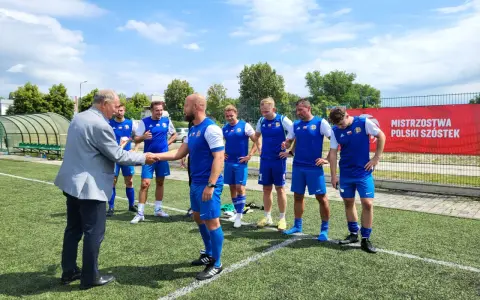 Starszy pan wita drużynę Grunwald Ruda Śląska na boisku. Piłkarze w niebieskich strojach czekają na rozpoczęcie meczu.