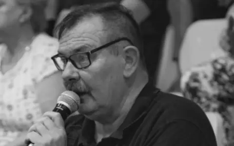 Krzysztof Janicki w okularach i z wąsami mówi do mikrofonu, w tle widać ludzi.