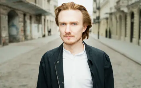 Bartosz Bednarski na tle miejskiej ulicy, z rudymi włosami i zielonymi oczami, w białej koszuli i ciemnej kurtce.