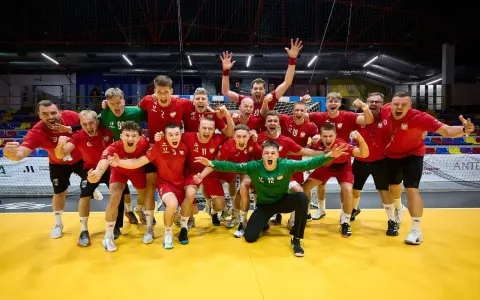 Drużyna piłkarzy ręcznych w czerwonych strojach świętuje zwycięstwo. Wśród nich Krzysztof Zimny, bramkarz w zielonym stroju, klęczy z wyciągniętymi rękami.