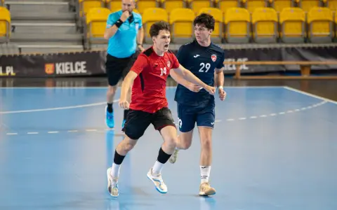 Jakub Smoliński w czerwonej koszulce z numerem 14 biegnie z piłką ręczną, obok zawodnik w granatowej koszulce z numerem 29.