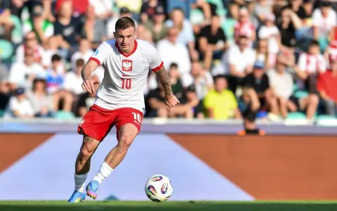 Jakub Kozłowski w akcji, w białej koszulce z orłem i czerwonych spodenkach, prowadzi piłkę na boisku.