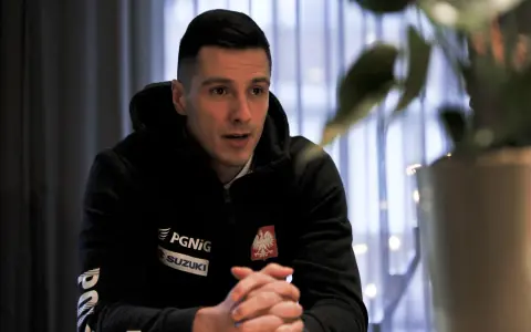 Maciej Pilitowski w czarnej bluzie z orłem i logiem Suzuki, z dłońmi złożonymi na stole, w tle zasłony i rośliny.