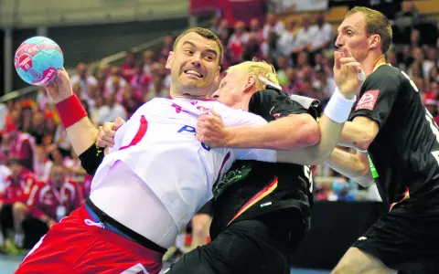 Gracz w białej koszulce z piłką ręczną próbuje wykonać rzut karny, podczas gdy przeciwnik próbuje go zatrzymać.