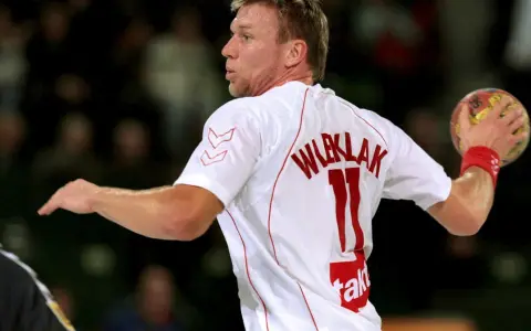 Damian Wleklak, numer 11, w białej koszulce z czerwonymi detalami, przygotowuje się do rzutu piłką ręczną.
