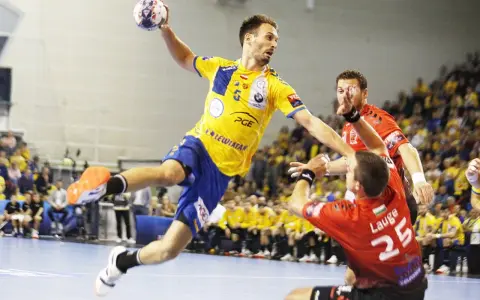 Piłkarz ręczny Telekom Veszprem w żółtej koszulce skacze do rzutu, broniony przez zawodnika w czerwonej.
