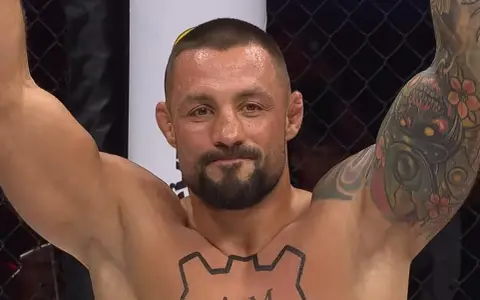 Mateusz Janur, wojownik MMA, z podniesionymi rękami i tatuażami na ramionach i klatce piersiowej, świętuje zwycięstwo.